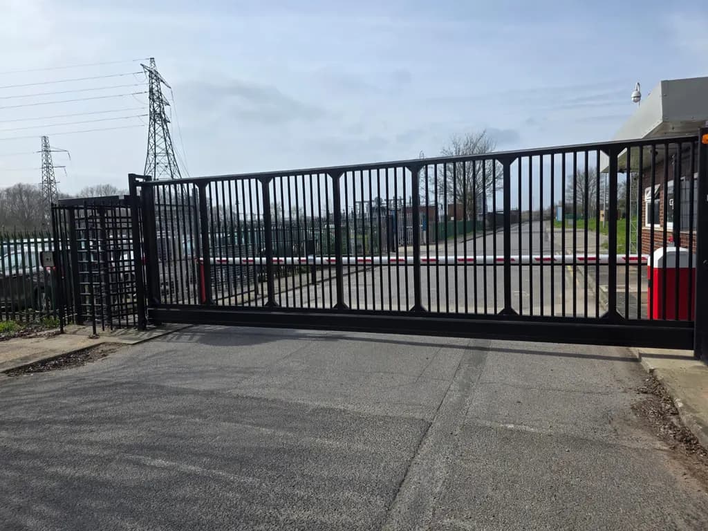 Large cantilever sliding gate closing an industrial access road, with a rising-arm barrier alongside
